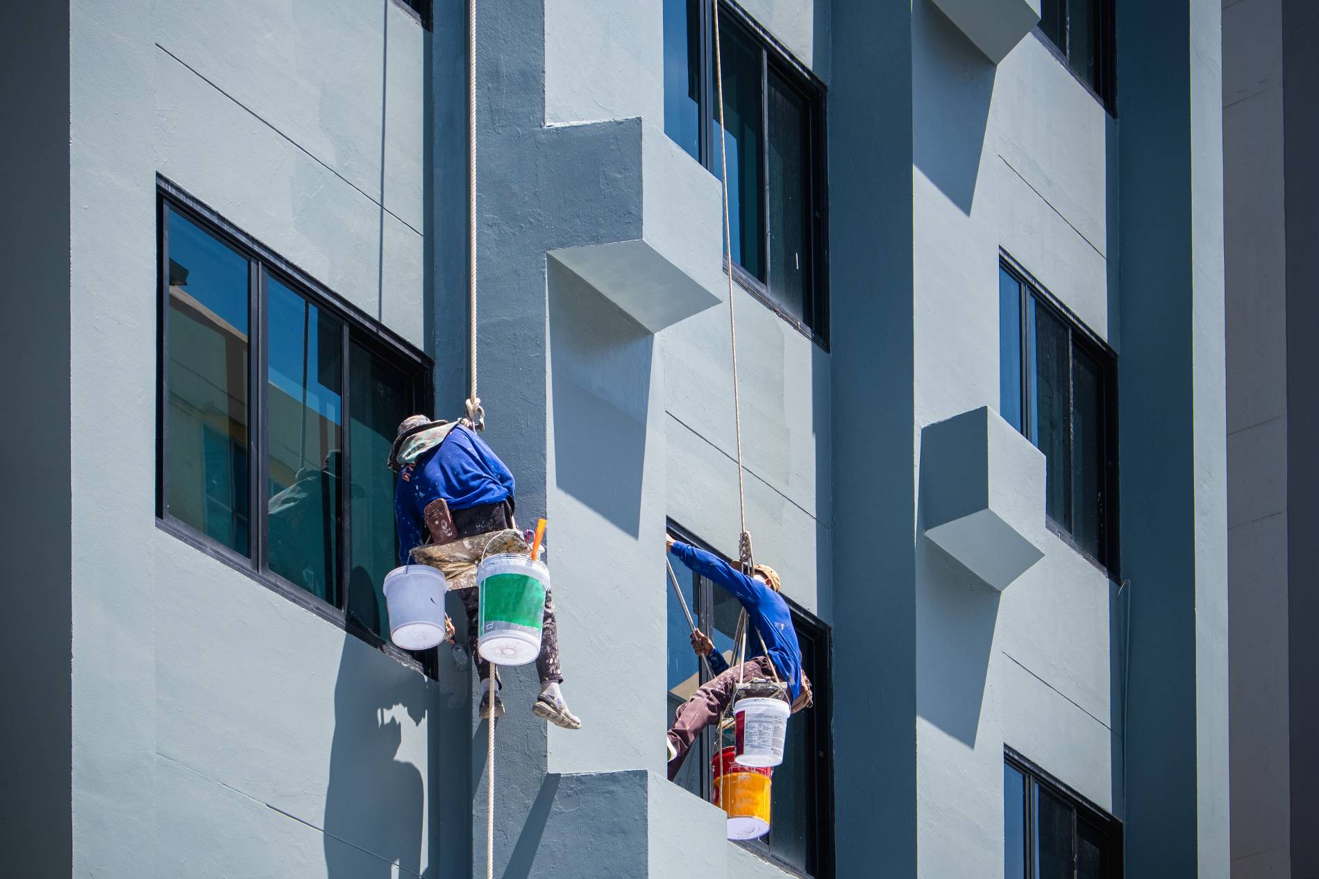 2 painters are hanging on ropes and harnesses paint the exterior of building.